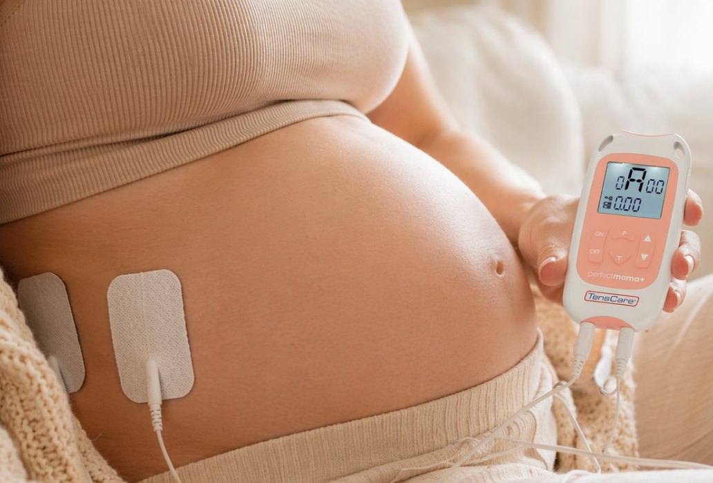 אישה בהריון עם אלקטרודות TENS על הבטן ומכשיר Perfect Mama+ ביד